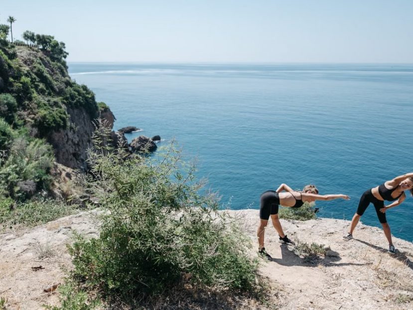 Two women stretching on a cliff by the ocean, representing fitness and outdoor exercise.