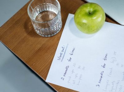A green apple, glass of water, and workout plan on a wooden desk symbolize healthy living.